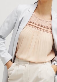 Persikofärgad blus med veck och spetsdetaljer vid halslinningen, matchad med en ljusgrå blazer och krämfärgade högmidjade byxor.