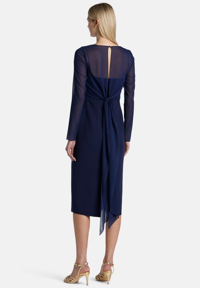 Tamsiai mėlyna suknelė su ilgomis permatomomis rankovėmis ir raktų skylutės nugarėle, surišta juosmenyje su plaukiančia juostele, iki kelių ilgio, lygaus tekstūros.