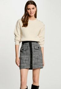 Cremfarbener Strickpullover mit geripptem Textur und dekorativen Knöpfen an der Schulter, kombiniert mit einem schwarz-weißen Tweedrock mit vorderem Knopfverschluss.