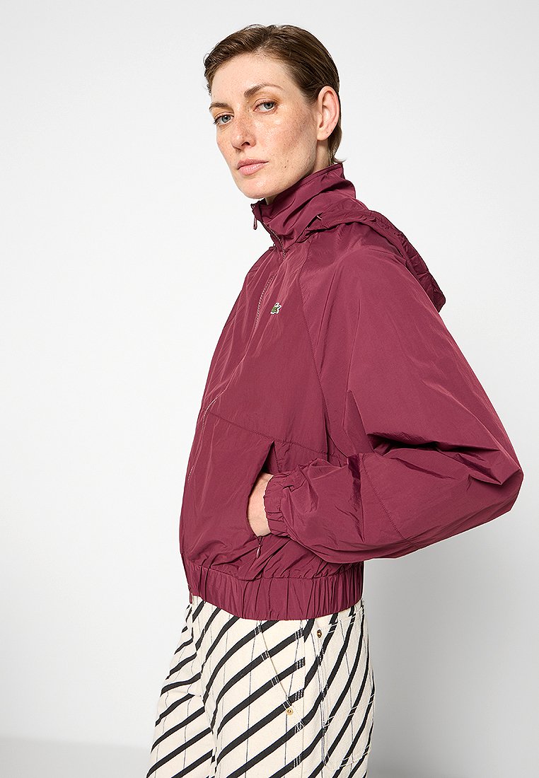 Bordeaux lichtgewicht jack met hoge kraag, elastische boorden en een voorzak. Gecombineerd met gestreepte offs-white en zwarte broek.