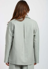 Donna con lunghi capelli castani che indossa una giacca oversized grigia chiara e pantaloni abbinati, vista di spalle su uno sfondo semplice.