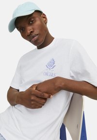 Camiseta de algodón blanca con bordado azul de un diseño floral y texto. Gorra azul clara. Modelo sentado en una silla con acentos azules.