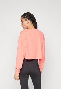 Kuttet langermet topp i lys korall, laget av lett stoff. Har senkede skuldre og en avslappet passform, sammen med svarte leggings.