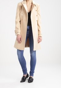 Béžový dvouřadý kabát se zlatými knoflíky, přiléhavý střih, nošený přes černý top, v kombinaci s modrými skinny džíny a černými mokasíny.