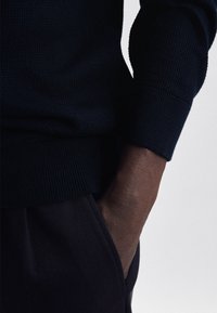Granatowy sweter teksturowany z prążkowanymi mankietami, noszony na ciemnych spodniach. Ręka spoczywa swobodnie w kieszeni, podkreślając kontrast materiałów.