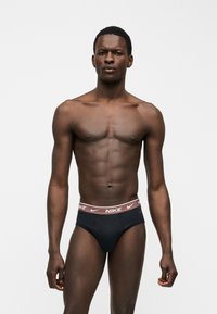 Μυώδης άνδρας όρθιος, φοράει μαύρα εσώρουχα Nike με ροζ ζώνη μέσης που φέρει λευκά λογότυπα Nike, σε απλό φωτεινό φόντο.