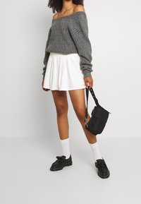 Grå stickad tröja med off-the-shoulder design, vit plisserad kjol, svart crossbody-väska, svarta skor och vita strumpor.