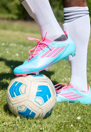 Botín de fútbol azul claro con acentos y cordones rosas, posicionado sobre un balón de fútbol con un patrón de detalles azules, en un campo de hierba.