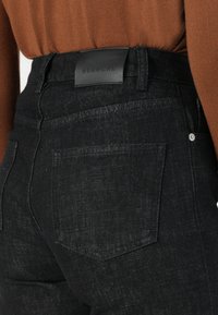 Κοντινό πλάνο από μαύρα τζιν με μια δερμάτινη ετικέτα "BLANCHE" στη μέση, συνδυασμένα με ένα καφέ πουλόβερ, αναδεικνύοντας στυλάτες, casual ενδυμασίες.