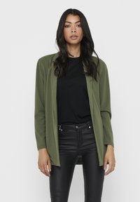 Blazer suave em verde oliva com mangas longas, design de frente aberta, sobre uma blusa preta, combinado com calças ajustadas pretas. Textura de tecido lisa.