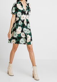 Blumenkleid mit schwarzem Hintergrund, das grüne und beige Blumen zeigt, kurze Ärmel und ein V-Ausschnitt-Design hat. Kombiniert mit glänzenden beige Ankle Boots.
