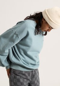Lichtblauwe sweatshirt met een ronde hals en een oversized pasvorm, gecombineerd met grijze geblokte broek. Gebreide crèmekleurige beanie op het hoofd.