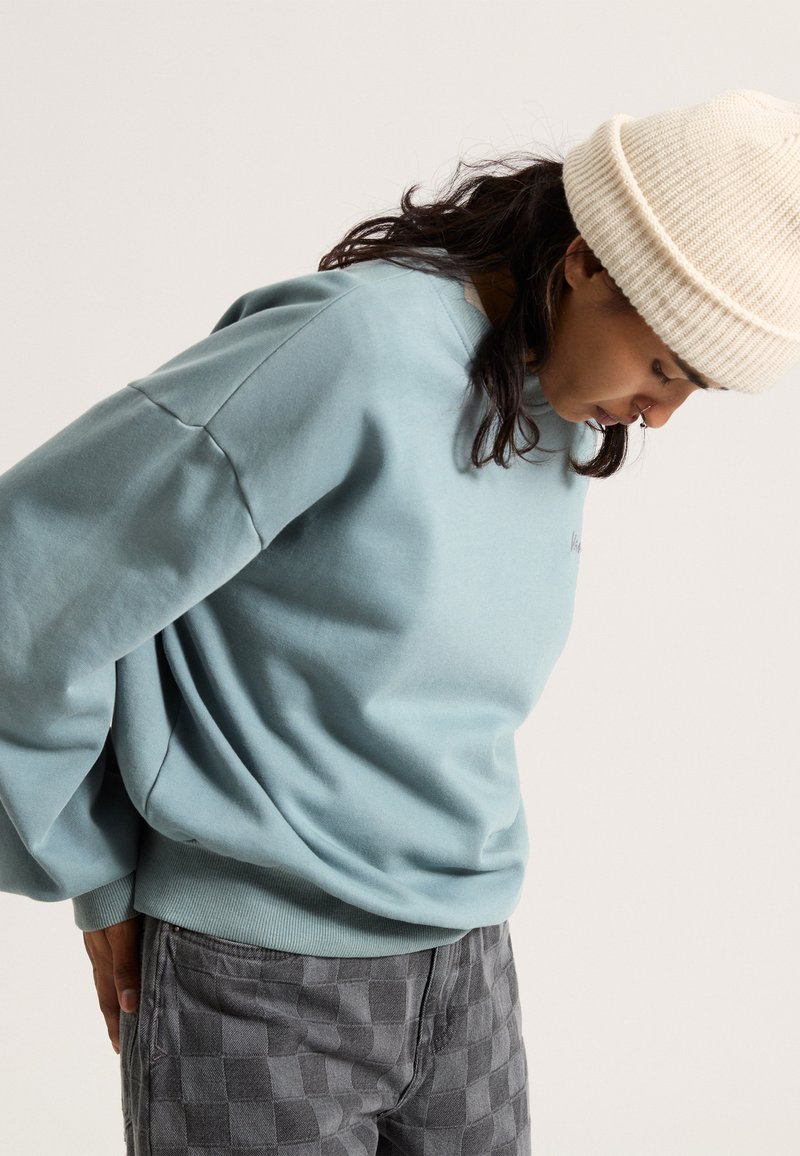 Himmelblauer Pullover mit Rundhalsausschnitt und oversized Schnitt, kombiniert mit grauen Karopants. Strickmütze in Cremefarbe auf dem Kopf getragen.
