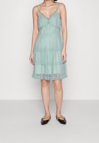 Světlý tyrkysový šaty s vrstveným designem, s ramínky na špagetách a krajkovými detaily. Textura je jemná s vrstveným volánkovým lemem.