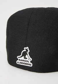 Black woven cap with a rounded shape, featuring a white kangaroo logo and "KANGOL J. LINDEBERG" text on a white background.