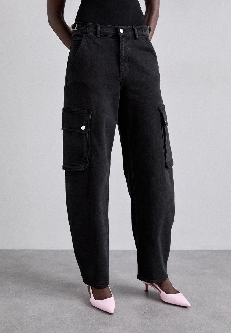 Persoană îmbrăcată cu pantaloni cargo negri, largi, cu buzunare laterale și pantofi slingback ascuțiți de culoare roz deschis, pe un podea gri.