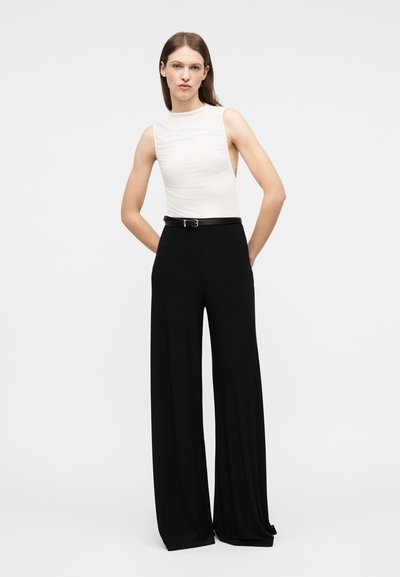 Γυναίκα με μακριά καστανά μαλλιά, φορώντας λευκό τοπ χωρίς μανίκια και μαύρο παντελόνι ψηλόμεσο με φαρδιά πόδια και μαύρη ζώνη, στέκεται μπροστά σε απλό φόντο.