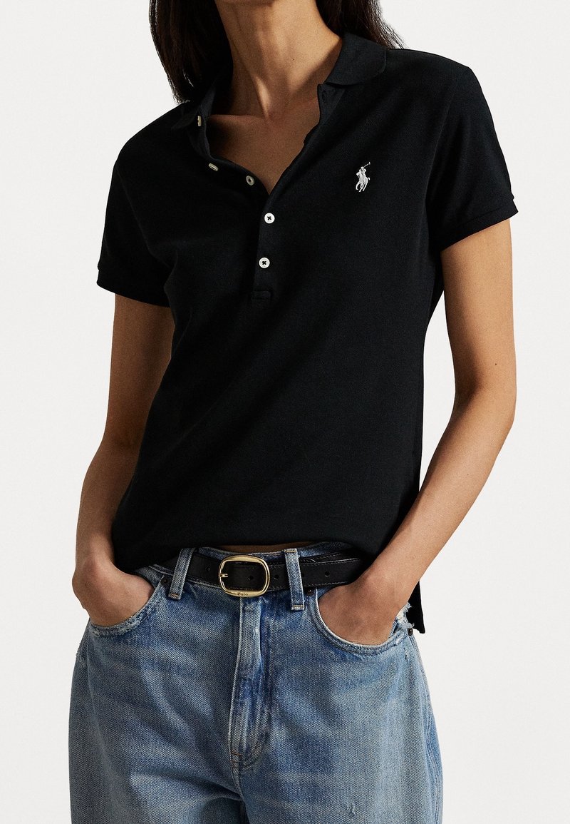 Femme portant un polo noir à manches courtes avec un logo blanc, rentré dans un jean bleu clair avec une ceinture noire, mains dans les poches.
