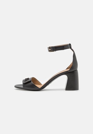 Sorte læder åbentået sandal med blokhæl og justerbar ankelrem med en guldbuckle på hvid baggrund.