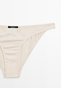 Beige thong underwear made of soft fabric with a smooth texture. Features a logo tag on the waistband and minimal stitching details.