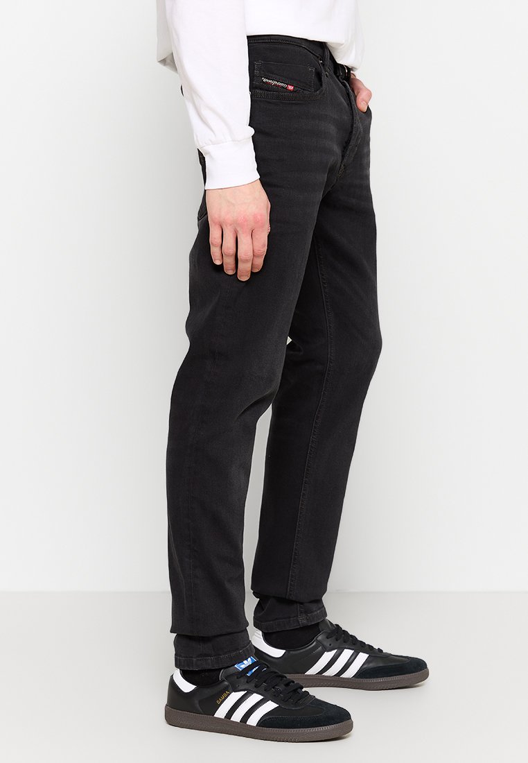 Jean en denim noir à coupe droite, cinq poches et logo discret au dos. Associé à des chaussures de sport noir et blanc.
