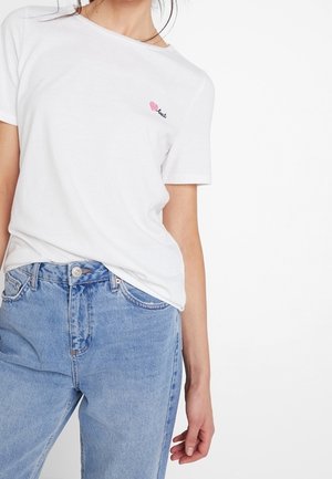 T-shirt en coton blanc à manches courtes, avec un petit logo en forme de cœur brodé en rose et noir sur la poitrine. Associé avec un jean bleu clair.
