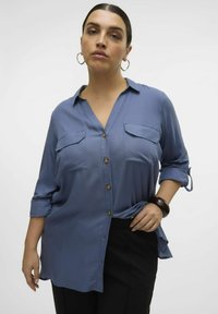 Blouse bleu clair à manches longues avec un devant boutonné, deux poches poitrine et une coupe ample. Présente des poignets retroussés et un ourlet arrondi.
