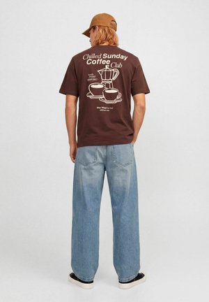Personne portant un t-shirt marron avec un graphisme de cafetière et de tasses ainsi que le texte "Chilled Sunday Coffee Club" dans le dos, jean bleu clair et casquette marron.