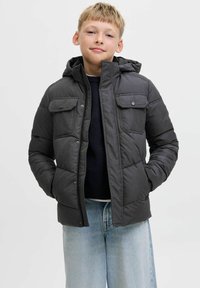 Vadderad jacka i mörkgrått tyg med dragkedja framtill, tryckknappsfickor och huva. Bärs över en marinblå tröja och ljusblå jeans.