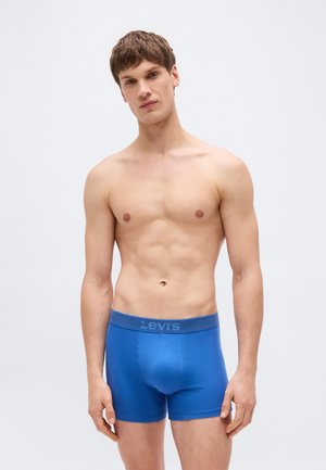 Jeune homme aux cheveux bruns courts portant un boxer Levi's bleu, debout devant un fond uniforme clair.