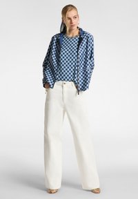 Giacca bomber a quadri blu con cappuccio, abbinata a una maglietta a fiori e pantaloni bianchi a gamba larga, completata da sandali nudi con cinturini.