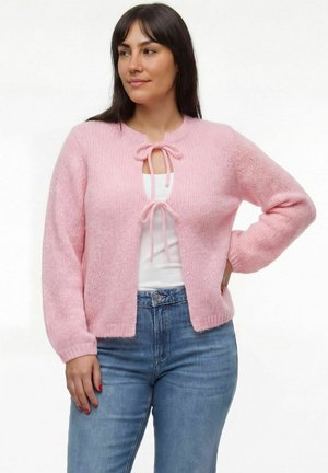 Nő világos rózsaszín kötött kardigánban, amelyet a nyaknál és a középső résznél kötött össze, fehér felsővel és kék farmerrel, egyik kezét a csípőjén tartva áll.