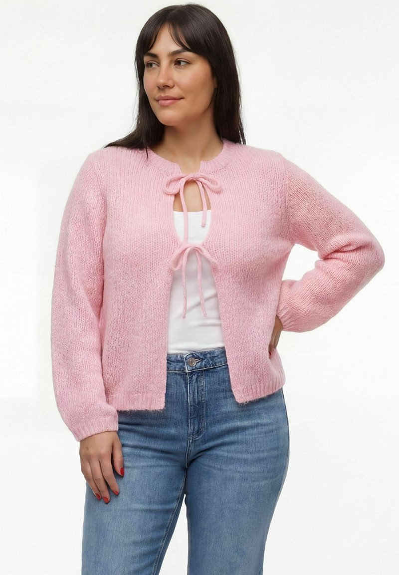 Sieviete valkā gaiši rozā dzīvokļu džemperi, sasietu pie kakla un vidukļa, ar baltu topu un zilām džinsām, stāv ar vienu roku uz gurna.