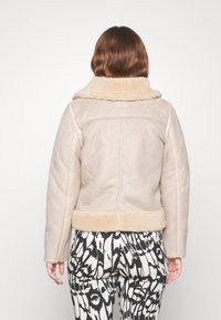 Žena stojící zády, oblečená v béžové kožešinové bundě a kalhotách s černobílým abstraktním vzorem na jednobarevném pozadí.