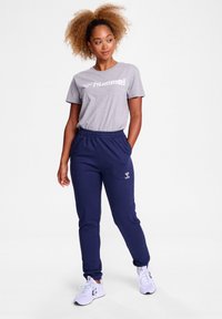 Grå T-shirt med hvidt logo, navy joggers med elastisk talje og mærkeaccent, parret med hvide træningssko. Glat tekstur, afslappet pasform.