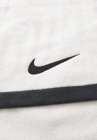 Fehér pamut törölköző, texturált felszínnel, fekete Nike logóval és sötétszürke vízszintes csíkkal az egyik szélén.