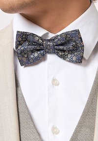 Un papillon floreale blu e oro realizzato in tessuto, con una forma classica e una finitura testurizzata, indossato con una camicia bianca e una giacca beige.