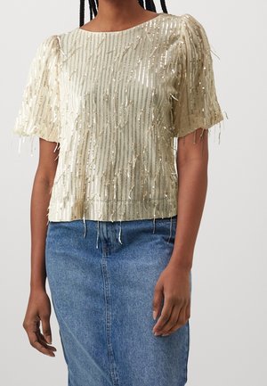 Kvinde iført en kortærmet beige top med lodrette gyldne palietter og et blåt denimskørt, stående mod en ensfarvet lys baggrund.
