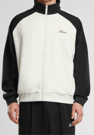 Homme portant une veste zippée noir et blanc avec des manches noires et le logo "Kani" sur la poitrine, ainsi qu'un pantalon noir.