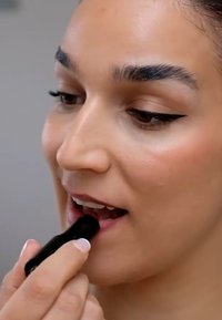 läppstift i en svart tub som appliceras på läpparna. Läpparna är en mjuk rosa färg och handen har en neutral manikyr. Slen hudstruktur.