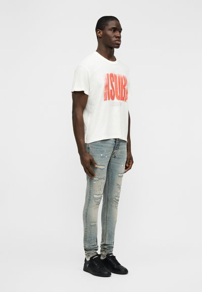 Λευκό μπλουζάκι με κόκκινο γραφικό κείμενο, συνδυασμένο με φθαρμένο στενό τζιν σε ανοιχτό μπλε χρώμα με σκισίματα, και μαύρα αθλητικά παπούτσια.