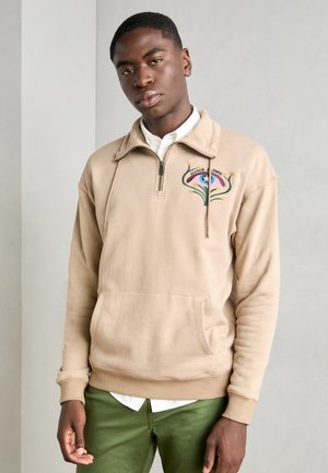 Béžová mikina na zips s vysokým golierom, zdobená farebnou vyšívanou aplikáciou na hrudi a prednou kapsou. Model má na sebe zelené nohavice.