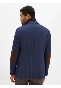 Marineblau kariertes Sakko mit braunen Ellbogenpatches, aus strukturiertem Stoff gefertigt. Mit Stehkragen und knöpfbaren Manschetten.