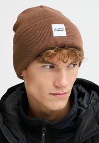 Smešani smeđi beanie kapu od rebrastog pletenog materijala, s bijelom etiketom s "NORTH BEND" logotipom. Nosi se s crnom jaknom.
