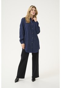 Chemise bleu marine à manches longues avec finition texturée et poche avant, associée à un pantalon noir à jambes larges et des chaussures à bouts pointus.