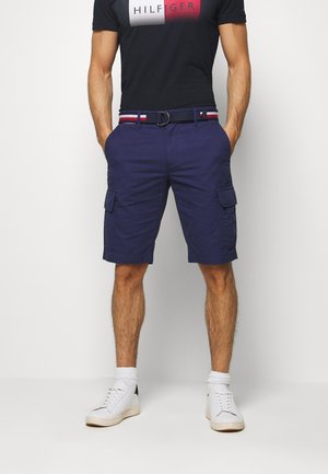 Hombre llevando pantalones cortos cargo azul marino, zapatillas blancas, calcetines blancos, camiseta negra de Tommy Hilfiger y un cinturón azul marino con rayas rojas, blancas y azules.