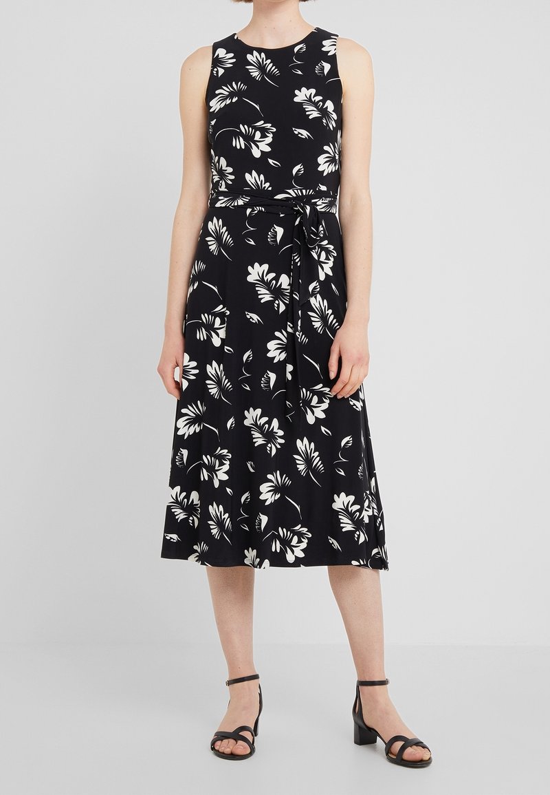 Černé midi šaty bez rukávů s bílým květinovým potiskem, s uvázaným páskem v pase, nošené s černými sandály na nízkém podpatku a sankou.