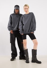 Felpe grigie in taglia oversize, abbinate a pantaloni neri strappati e stivali da motociclista neri. La donna indossa pantaloni corti neri da motociclista.