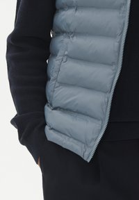 Gai pelēks pūkains vests ar quiltinga dizainu, ar rāvējslēdzēju, un tumši zila džemperis zem tā, kuram ir ribota adījuma tekstūra uz piedurknēm.