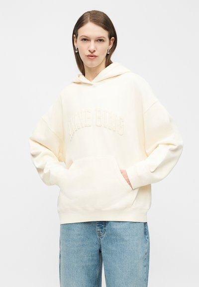 Jeune femme portant un sweat à capuche crème avec les mains dans la poche avant et un jean bleu clair, debout devant un fond uni.